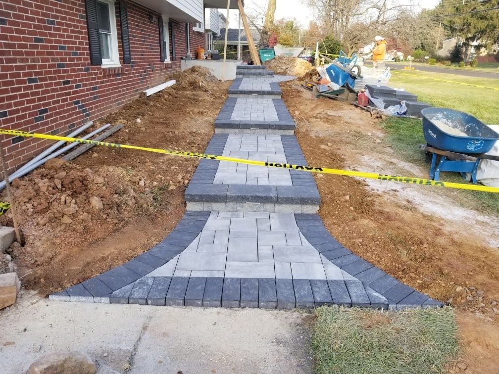 Brick walkway under construction beside a house, with gray pavers, dirt trenches, and caution tape
