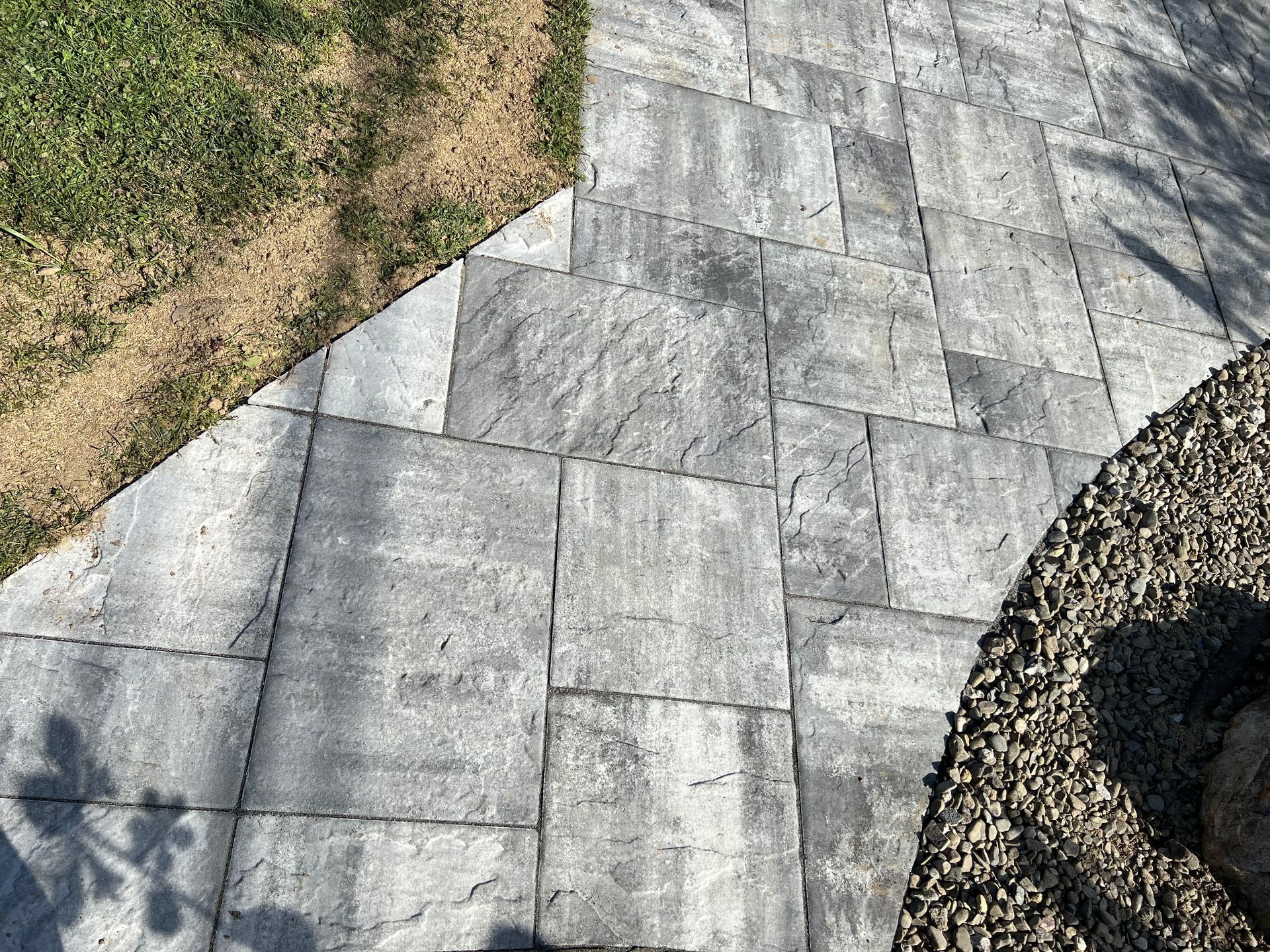 Gray stone paver walkway beside grass and gravel landscaping, with tree shadows.