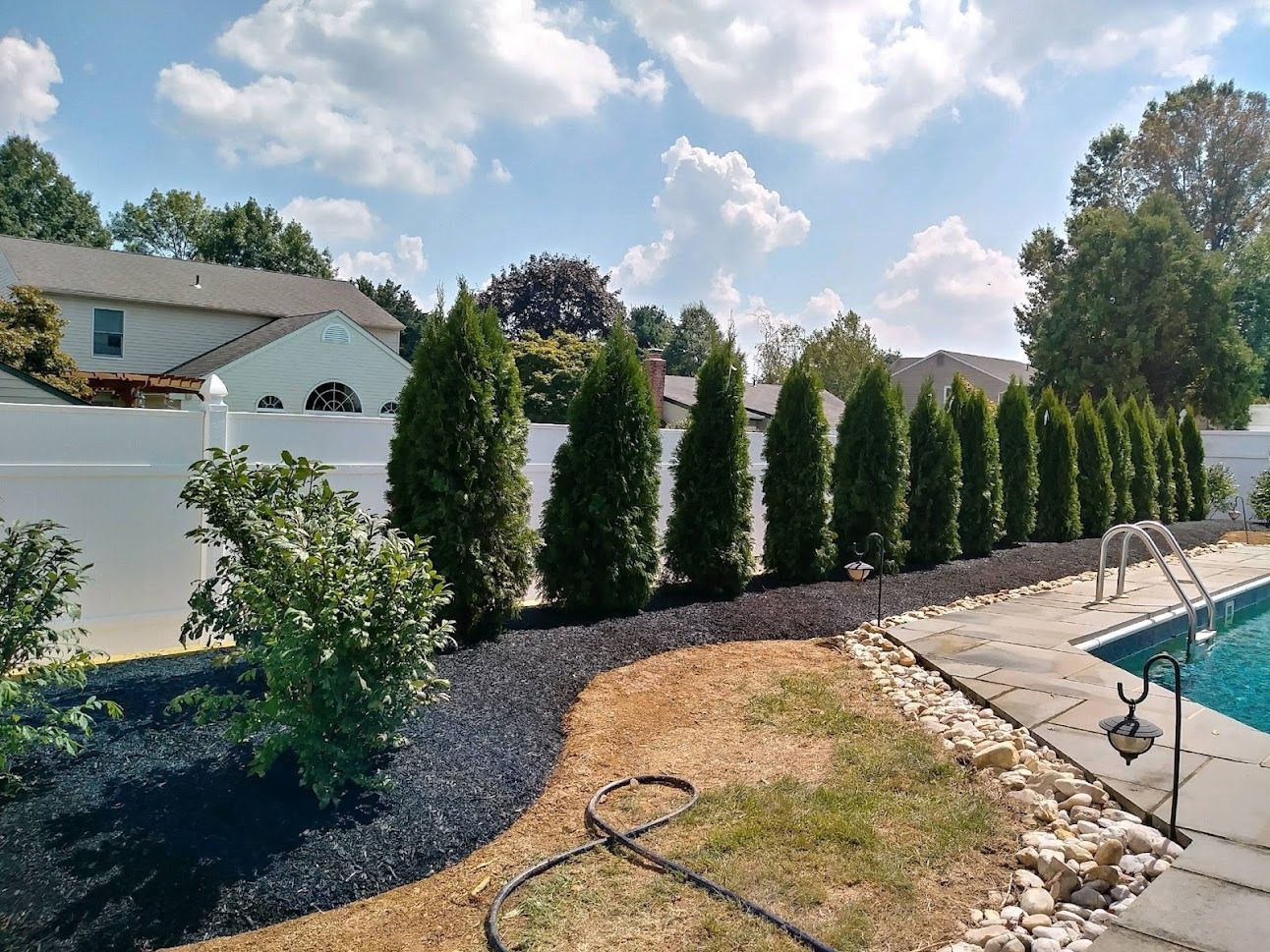 Backyard with tall evergreen hedges, a pool, and a stone patio under a bright cloudy sky