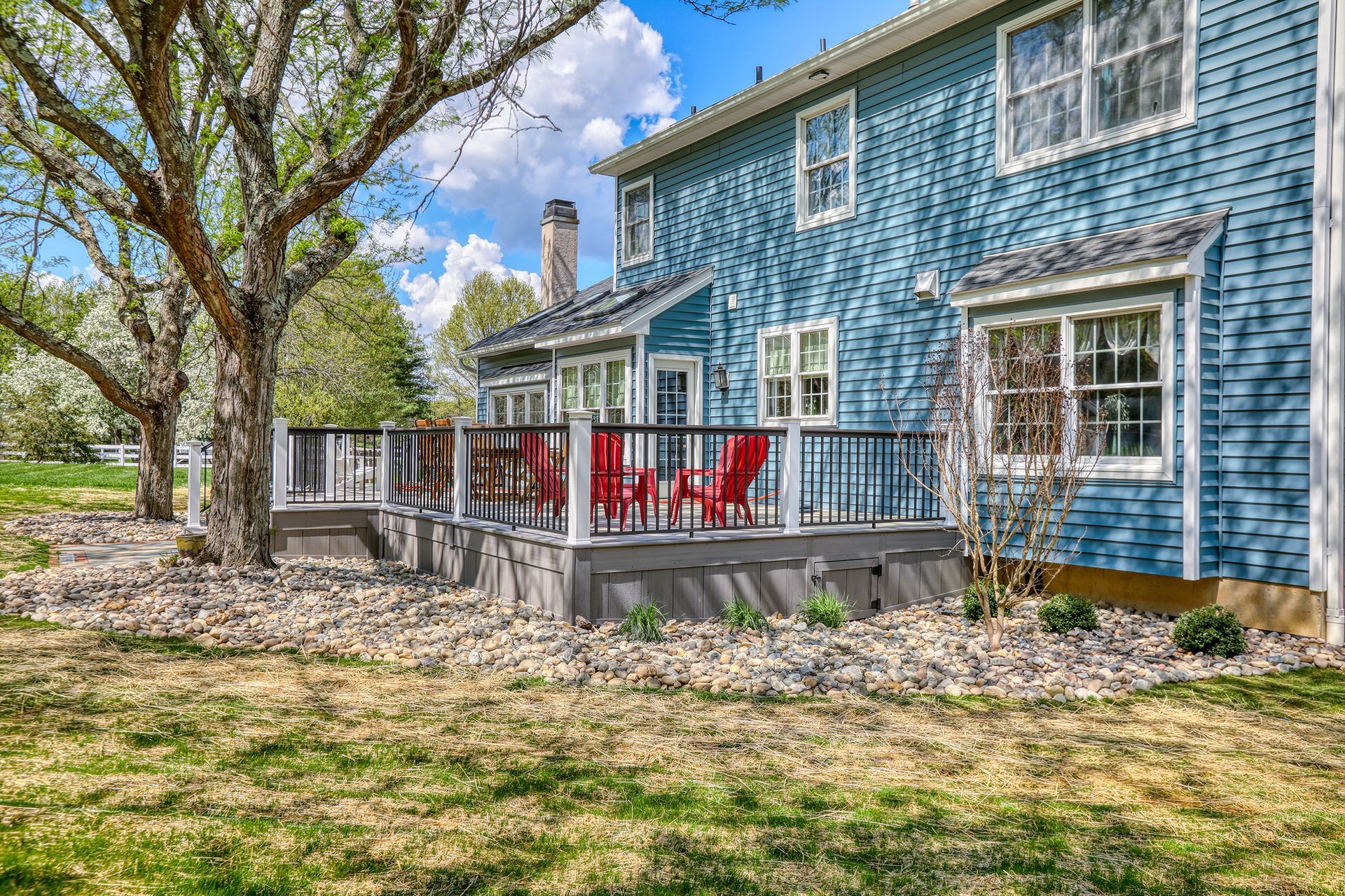 Blue two-story house with a small deck and red chairs under leafy trees in a yard
