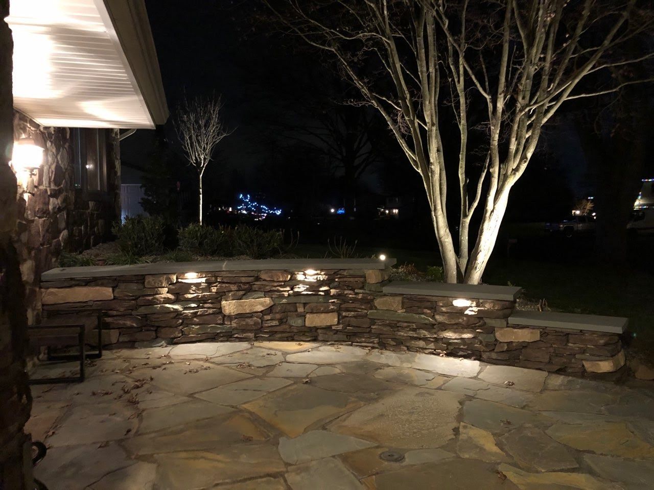 Stone patio with low retaining wall and bare trees lit at night beside a house porch