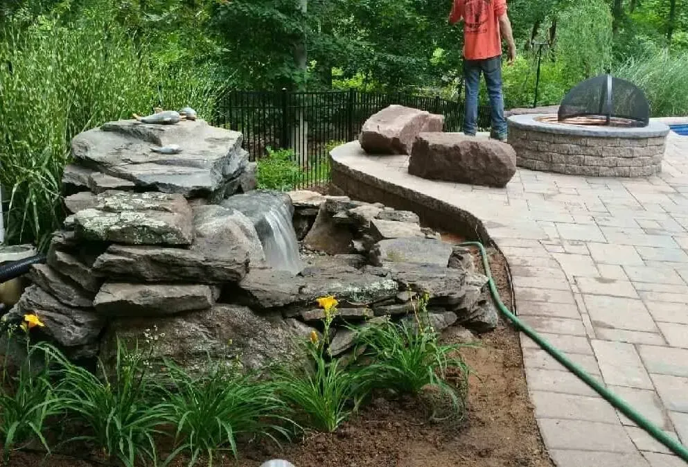 Waterfall feature with rocks and plants next to a patio and a fire pit. A person is standing near the fire pit.