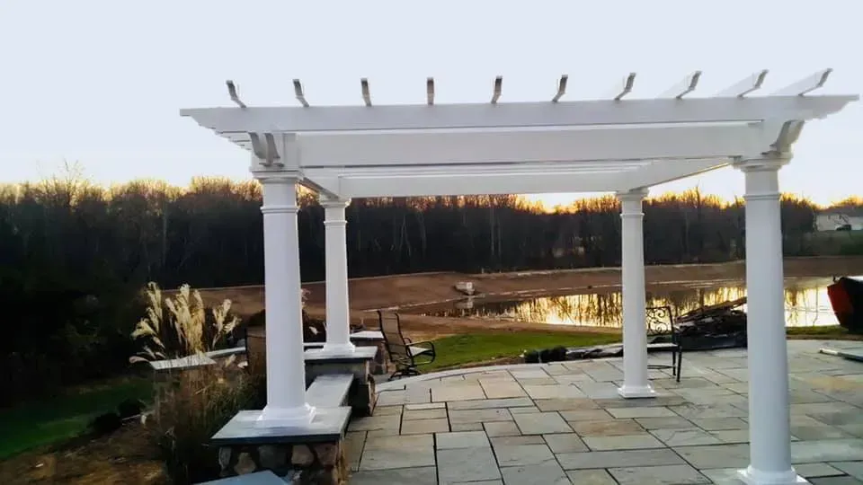 White pergola over a stone patio overlooking a lake and trees at dusk.
