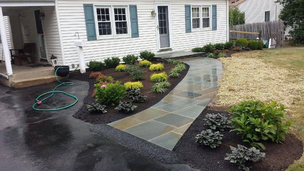 Front yard landscaping: stone path, flower beds with mulch, house with blue shutters, wet asphalt driveway.