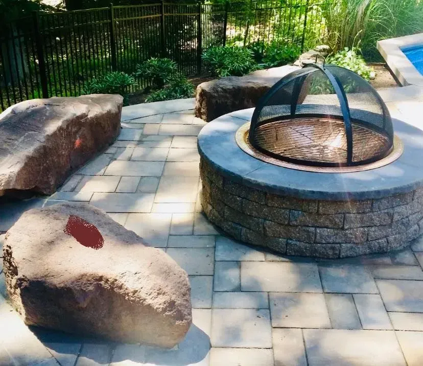 Fire pit with rocks for seating on a stone patio near a pool and greenery.