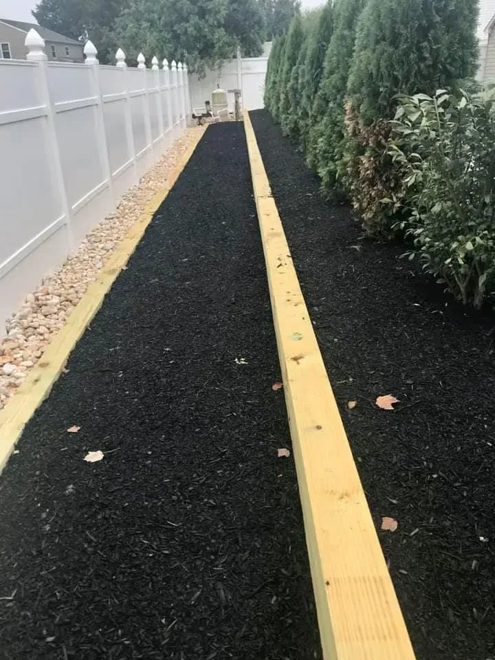 A narrow backyard path with black mulch, bordered by a wooden beam and a white fence.