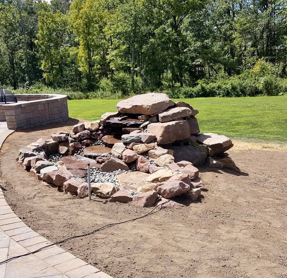 Rock waterfall feature in a yard, with a paved patio and surrounding grass, next to a retaining wall.