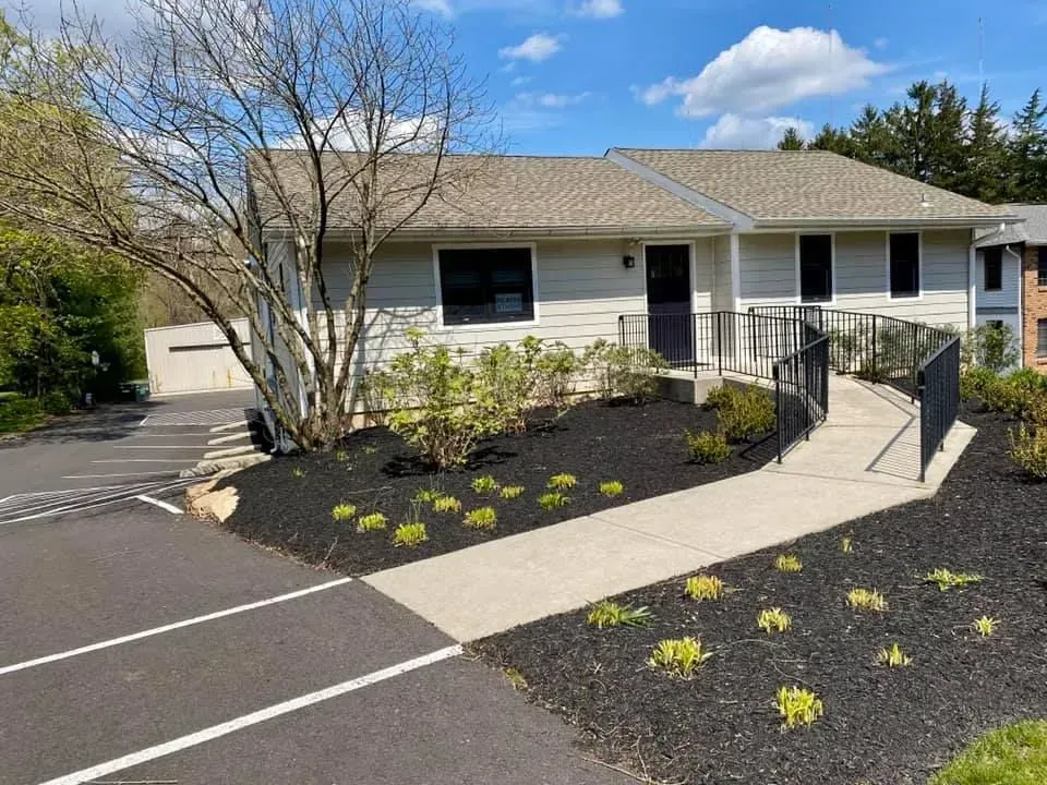 Exterior of a light-colored, single-story building with a concrete walkway leading to the entrance. Dark mulch and landscaping are in the foreground.