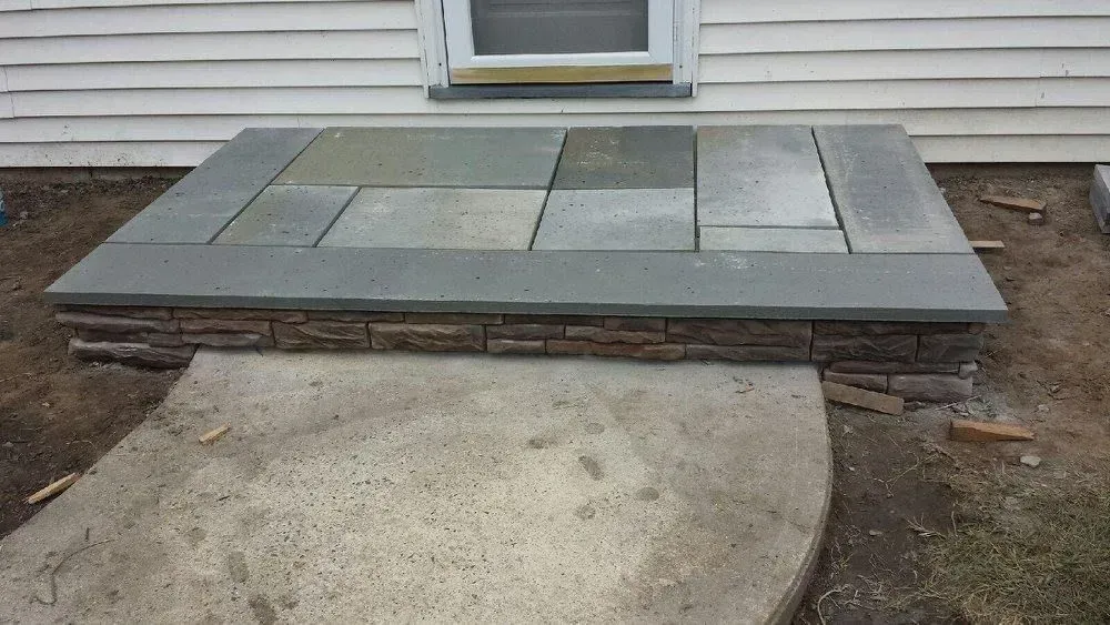 Stone-tiled stoop with a raised border and a brick-like facade, in front of a window and a concrete walkway.