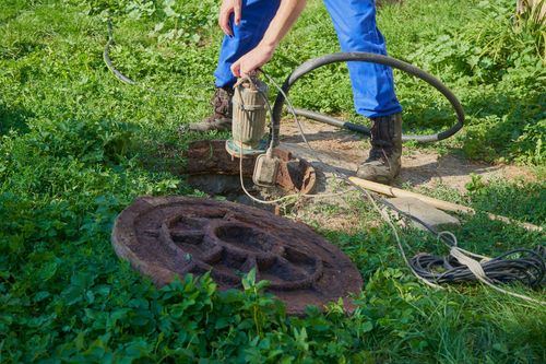 Worker lifting a rusty manhole cover beside a hose on a grassy path.