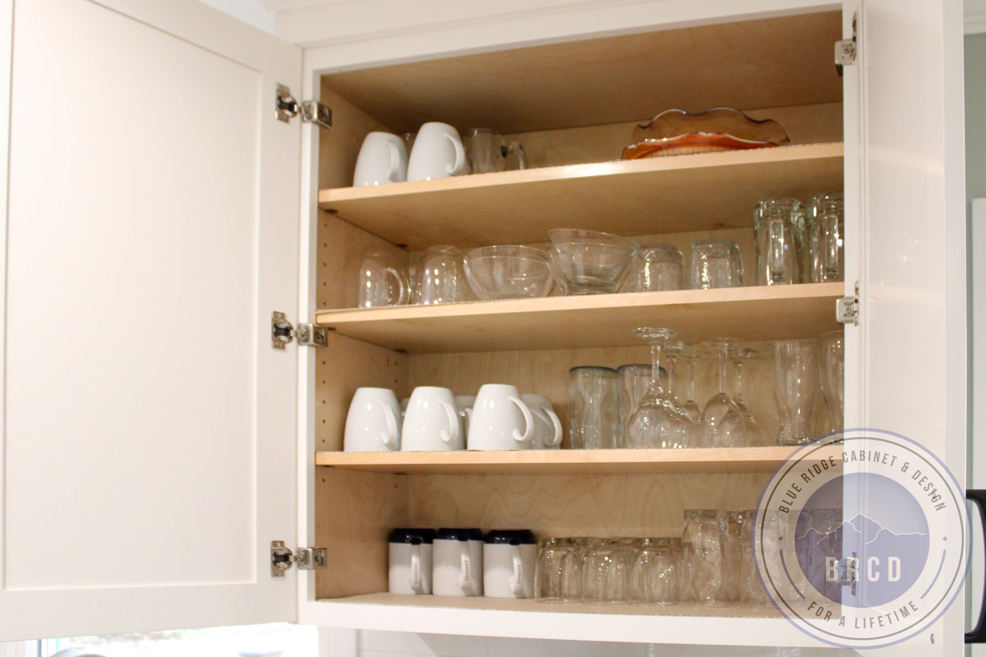 Open kitchen cabinet with neatly arranged white mugs, glassware, and bowls on wooden shelves