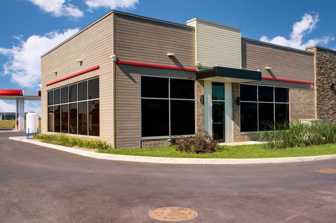Empty commercial building with tan siding, large dark windows, and red trim on a paved lot