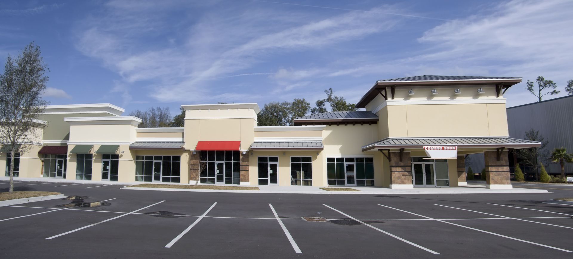 A strip mall with beige walls, a red awning, and a mostly empty parking lot under a blue sky.