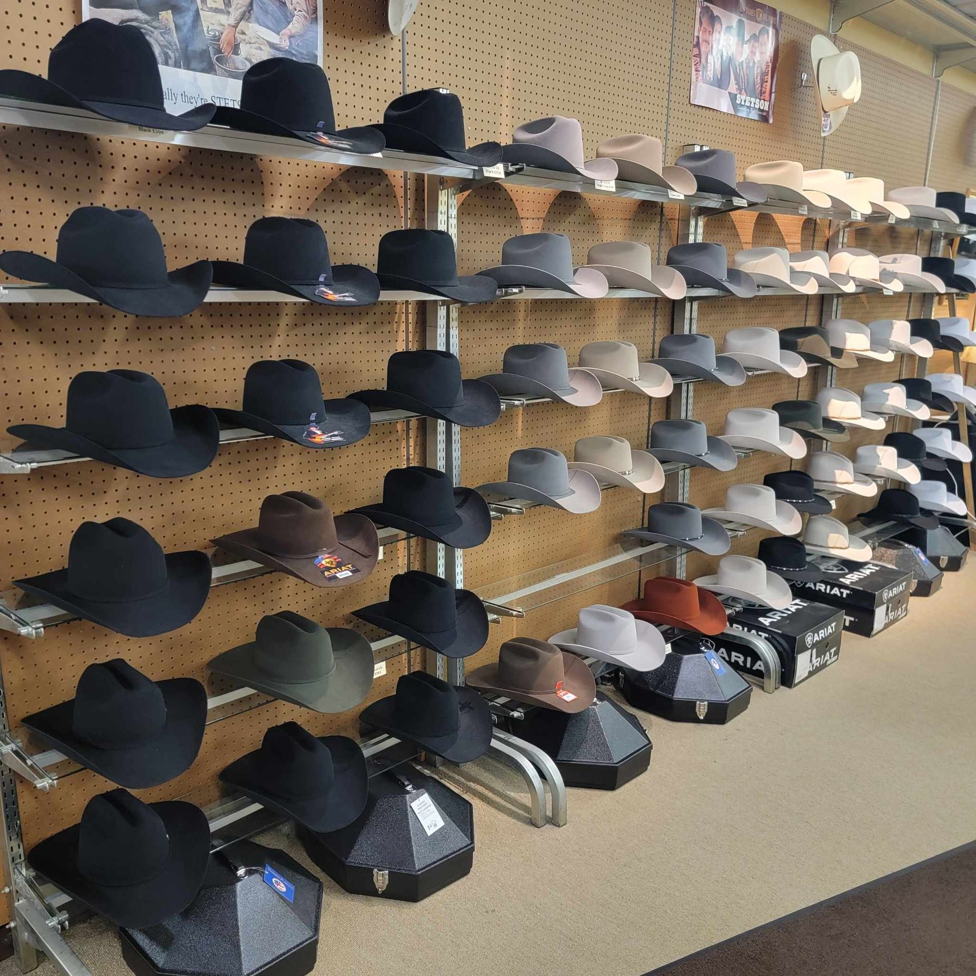A retail display of numerous cowboy hats in black, grey, and white, arranged on multiple shelves against a pegboard wall.