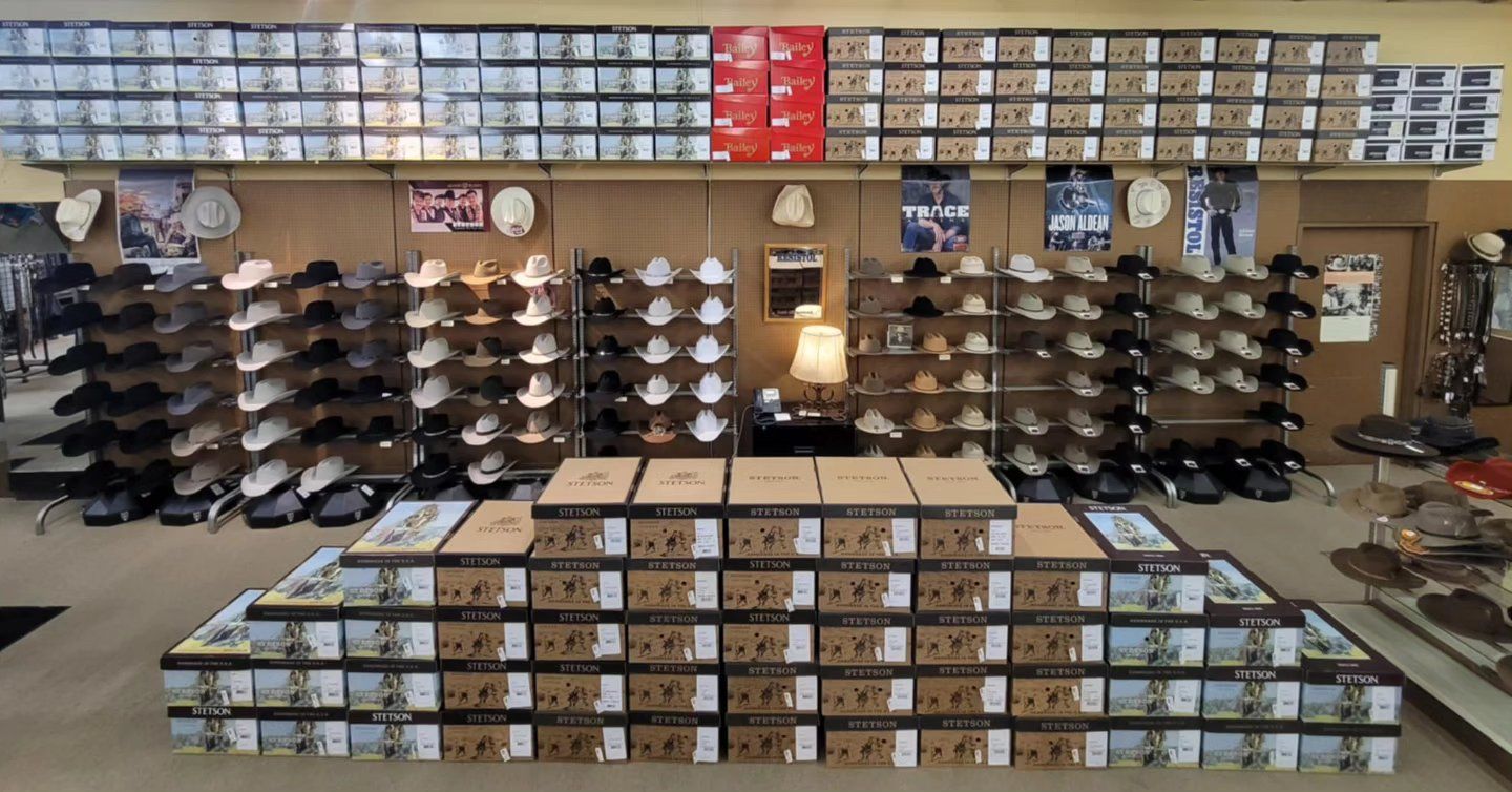 A retail store aisle featuring shelves stocked with western hats and a large, multi-level pyramid display of boot boxes.