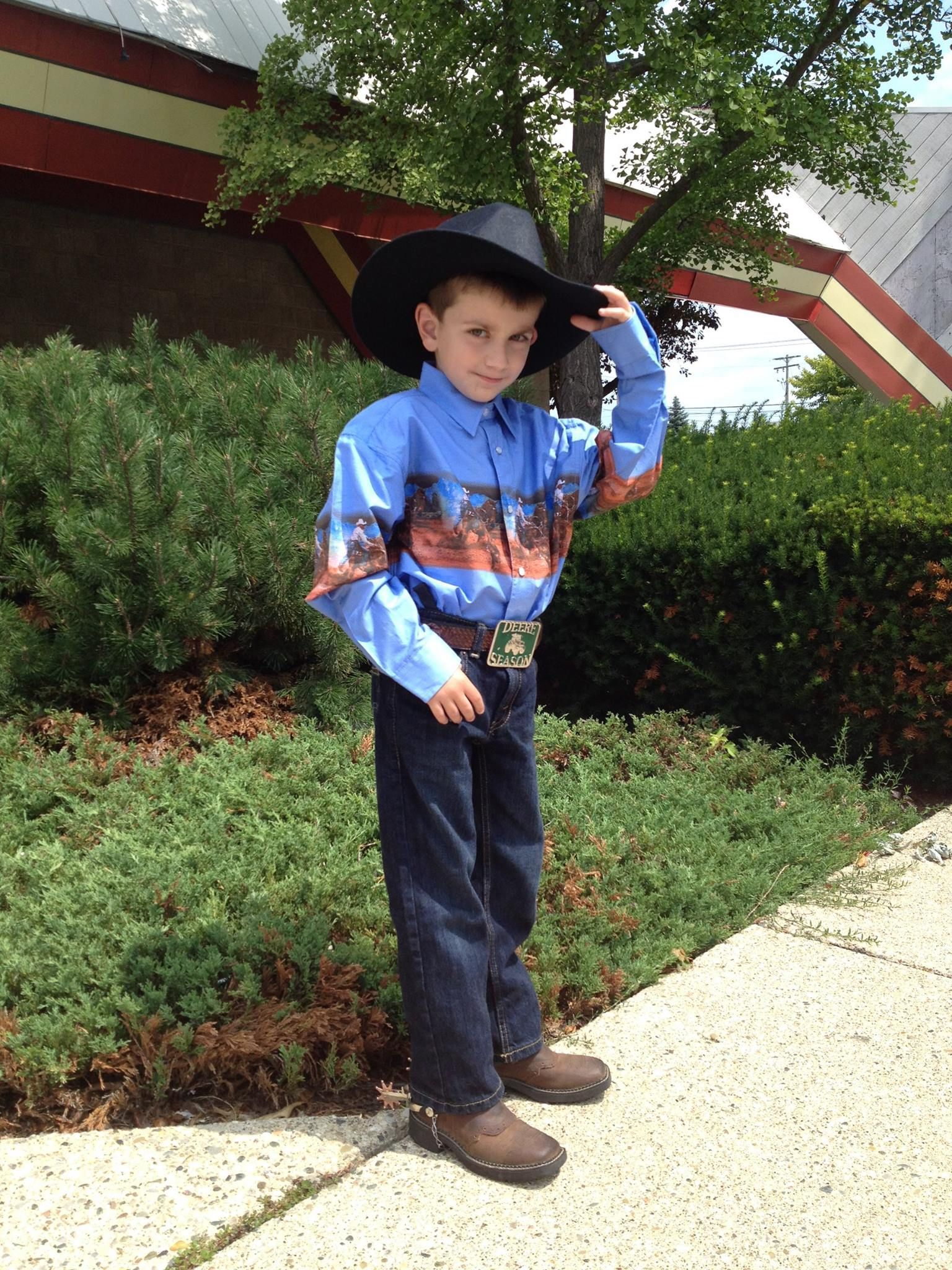 A person in a cowboy hat, patterned blue shirt, and jeans stands on a sidewalk in front of bushes and a building.