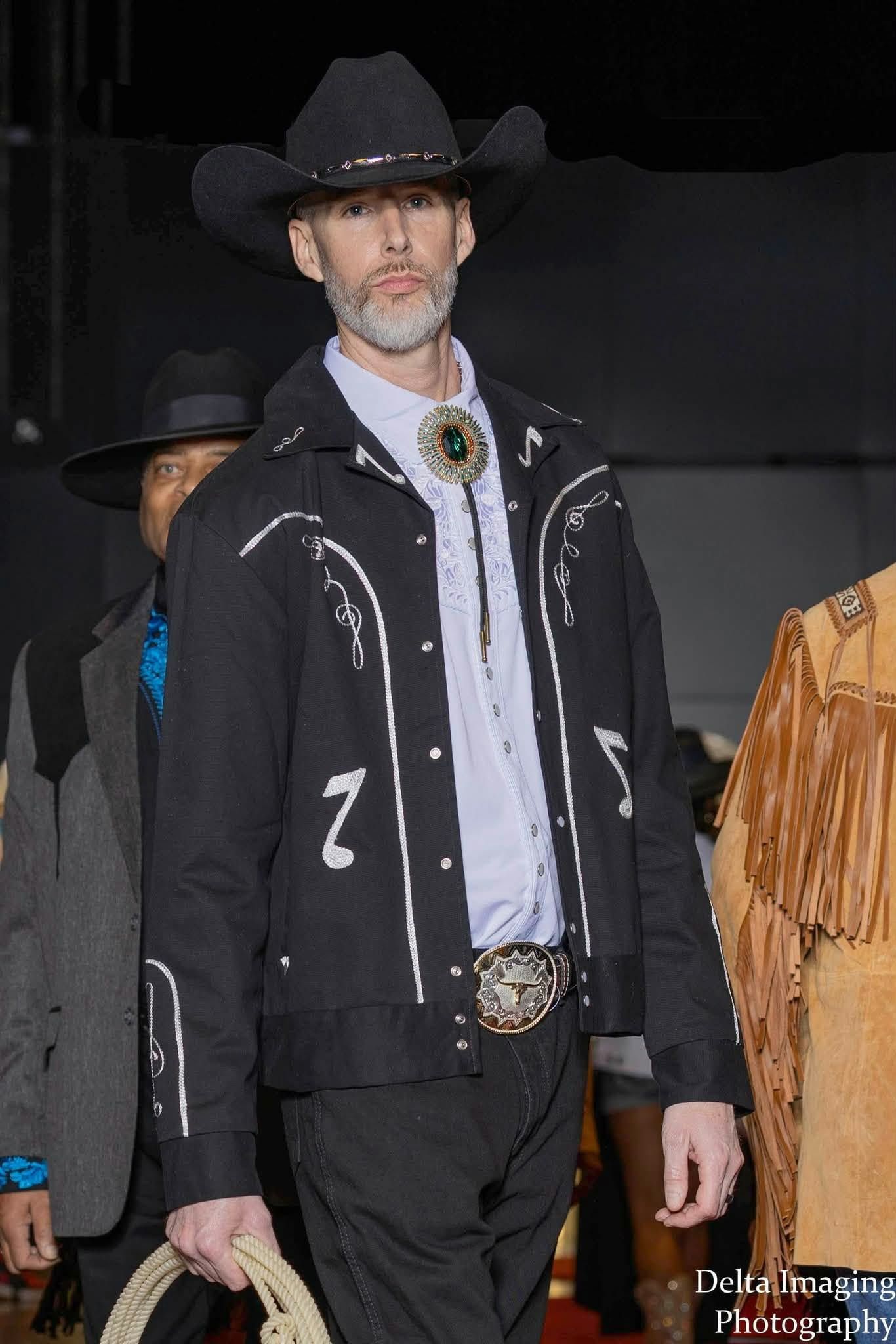 A person models a black cowboy jacket with silver accents, a bolo tie, and a hat while walking on a runway.