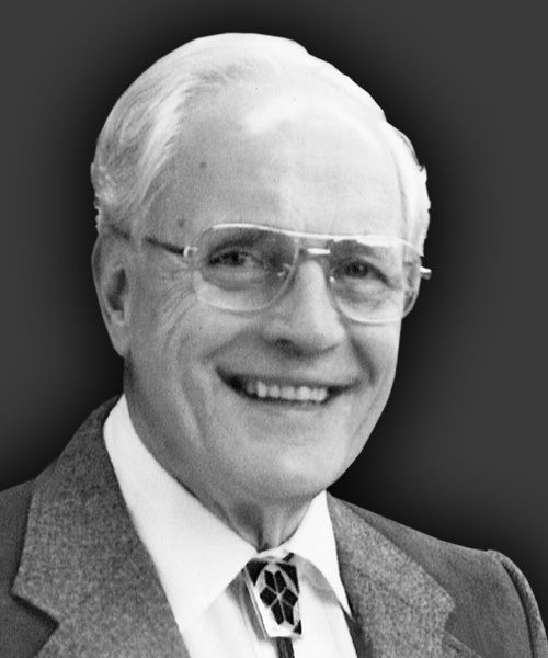 A head-and-shoulders portrait of a smiling person wearing glasses and a bolo tie, set against a dark background.