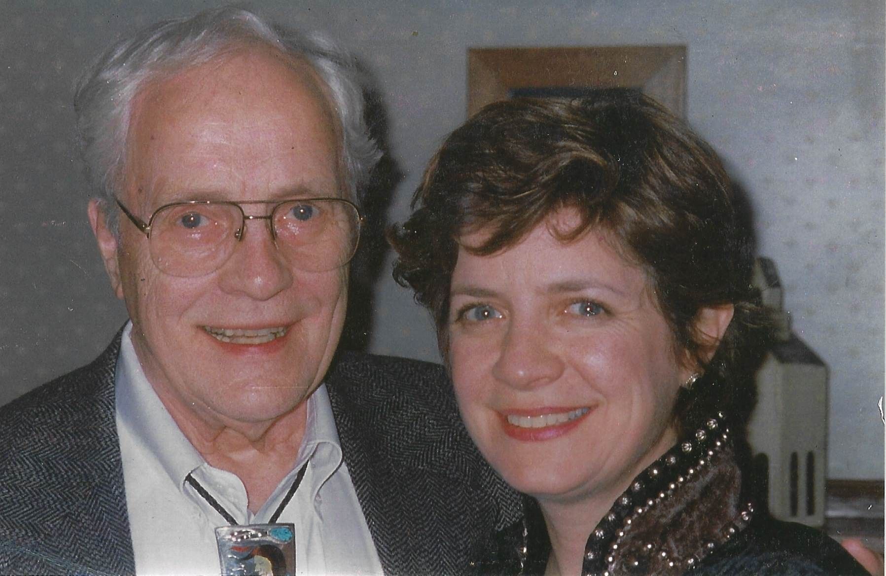 Two people smiling side-by-side. The man wears glasses and a bolo tie; the woman wears a dark collared top.