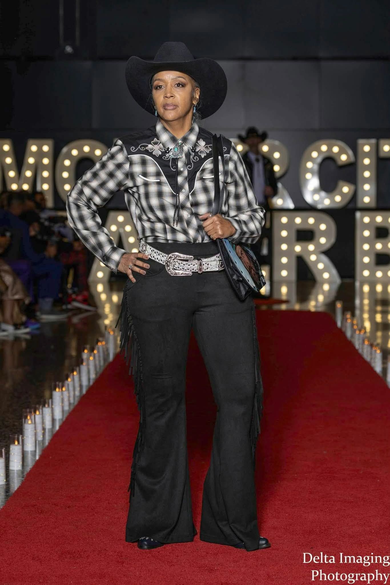 A model in a black cowboy hat and plaid Western shirt walks down a red carpet lined with lights in a studio.