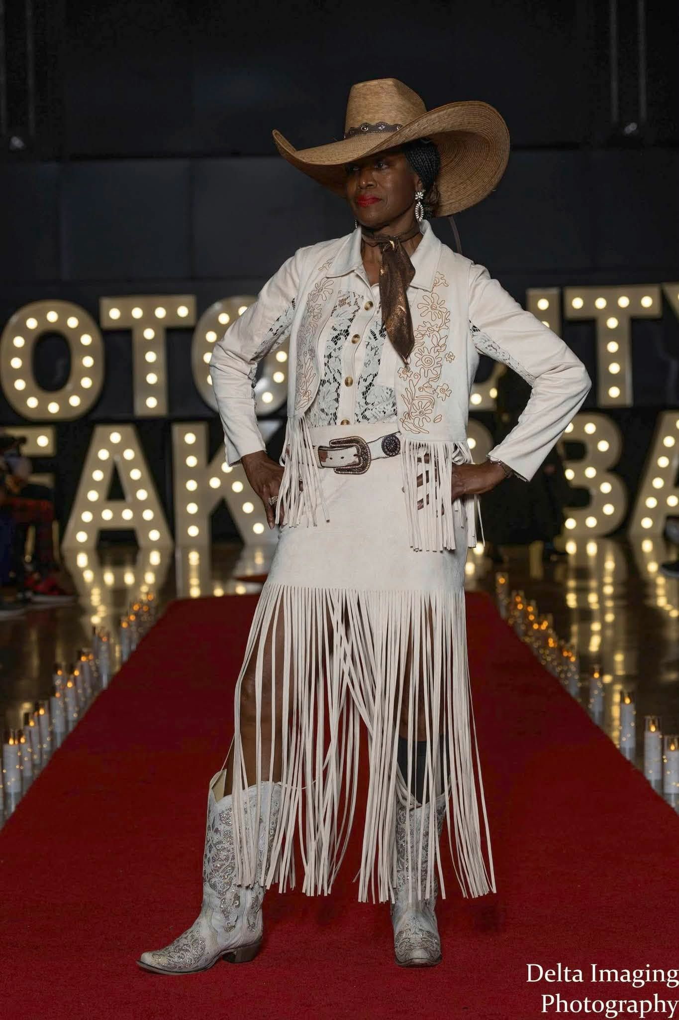 A model in a straw cowboy hat, patterned jacket, and fringed skirt walks a red carpet before a lit marquee.
