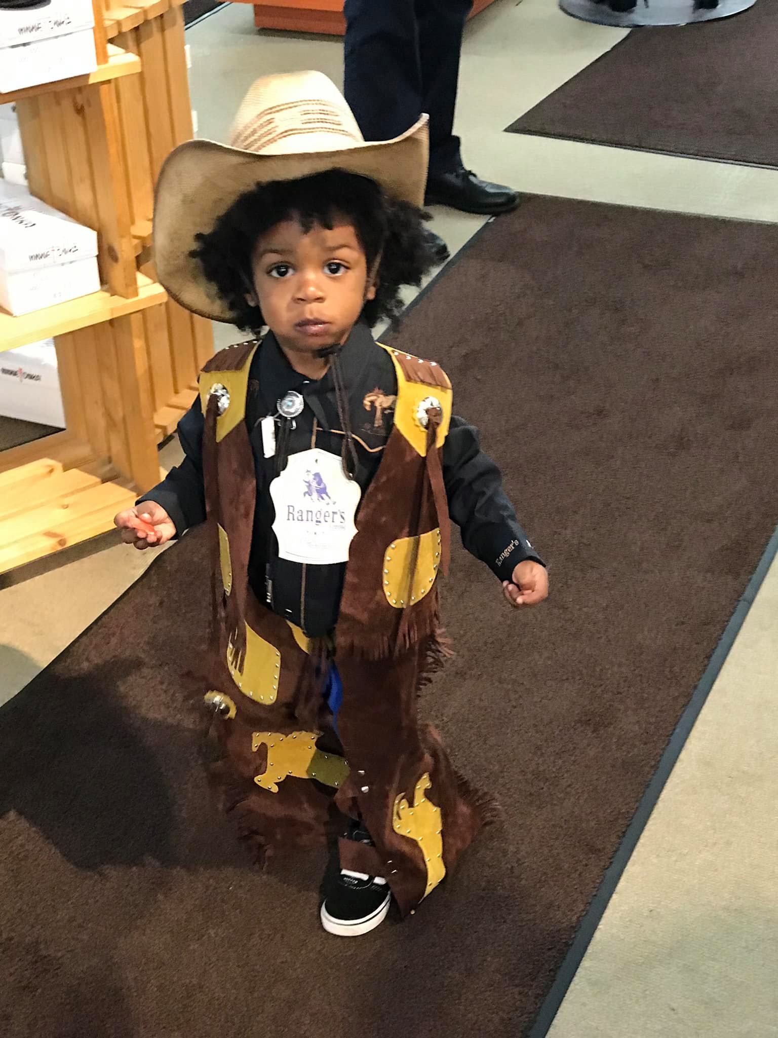 A child wearing a cowboy costume, including a hat and fringed chaps, standing on a brown rug in a store.