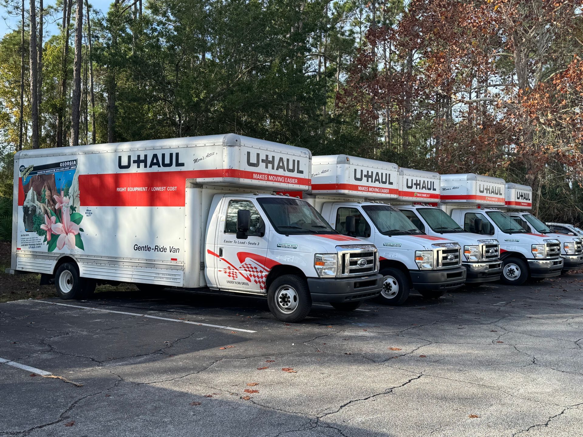Four U-Haul moving trucks parked in a lot, lined up beside trees in autumn.