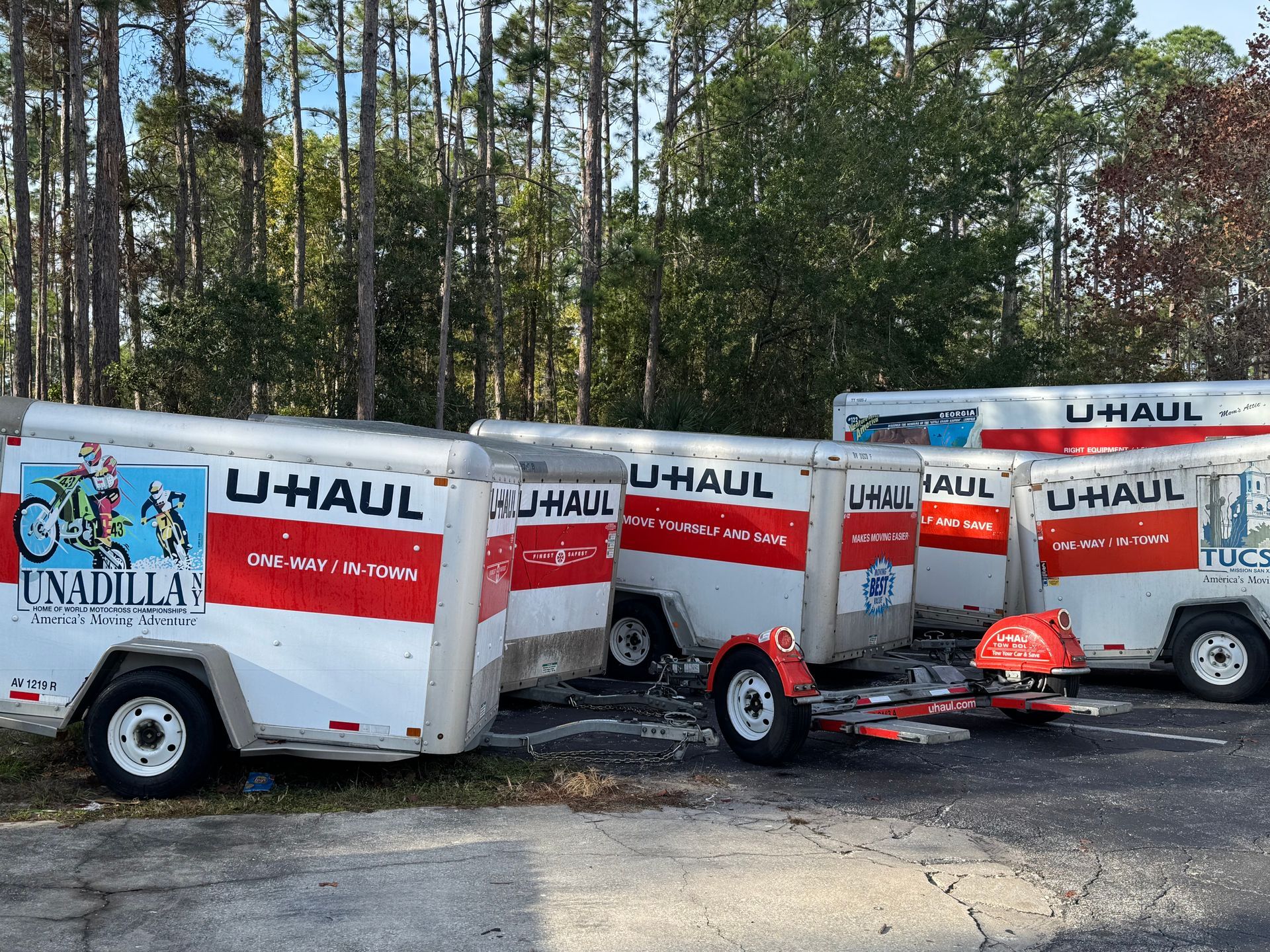 U-Haul trailers and a tow dolly lined up in a wooded lot, with a truck in the background.