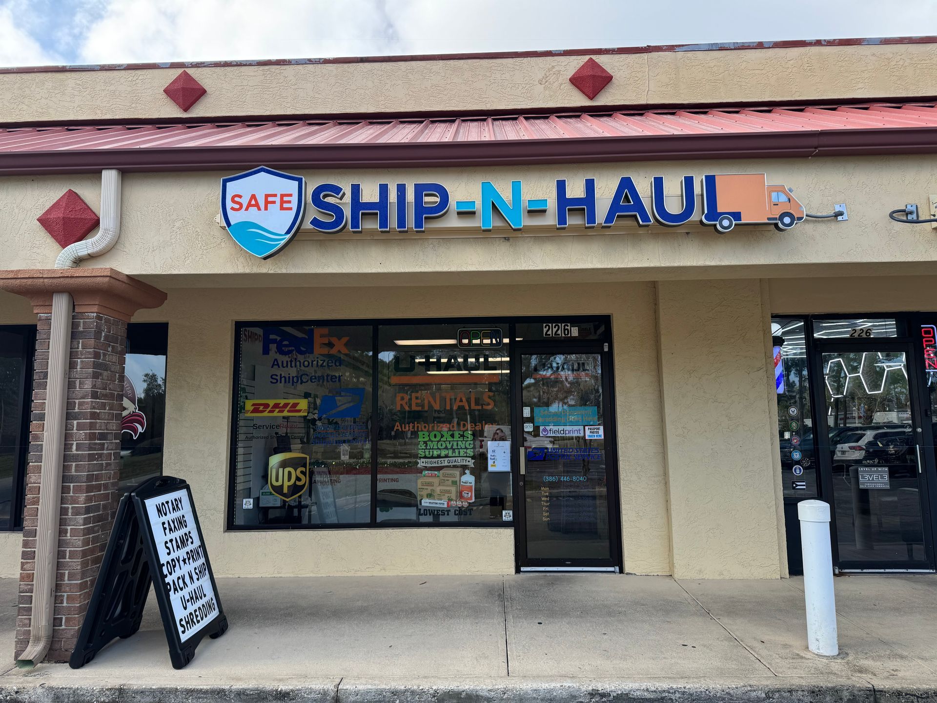 Strip mall storefront for SAFE Ship-N-Haul with blue and orange sign, sidewalk sign, and glass entrance.