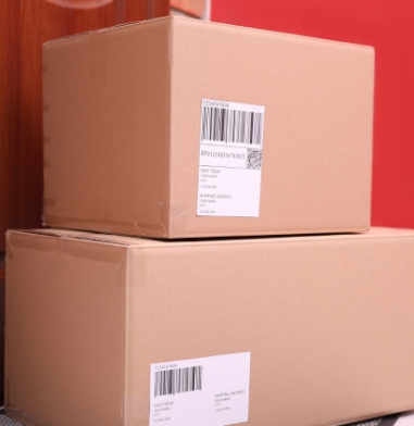 Two cardboard shipping boxes stacked against a red wall, each with a white barcode label.