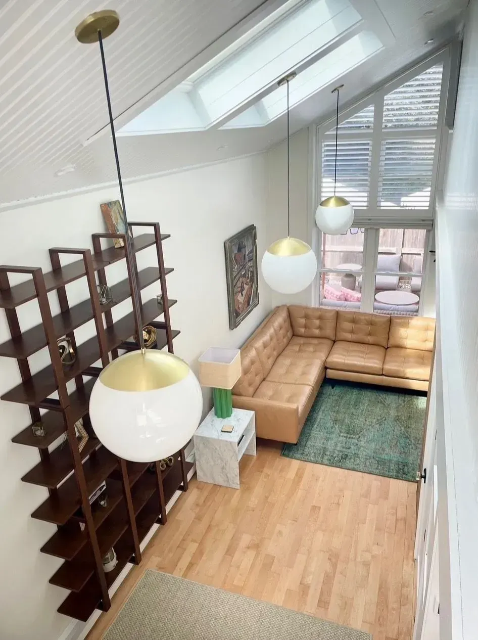 Bright loft living room with tan sectional, green rug, hanging globe lights, skylight, and tall shelving divider