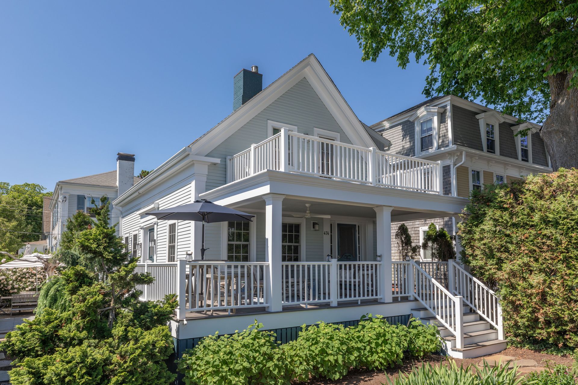 White house with wraparound porch, white railings, and green landscaping under a blue sky