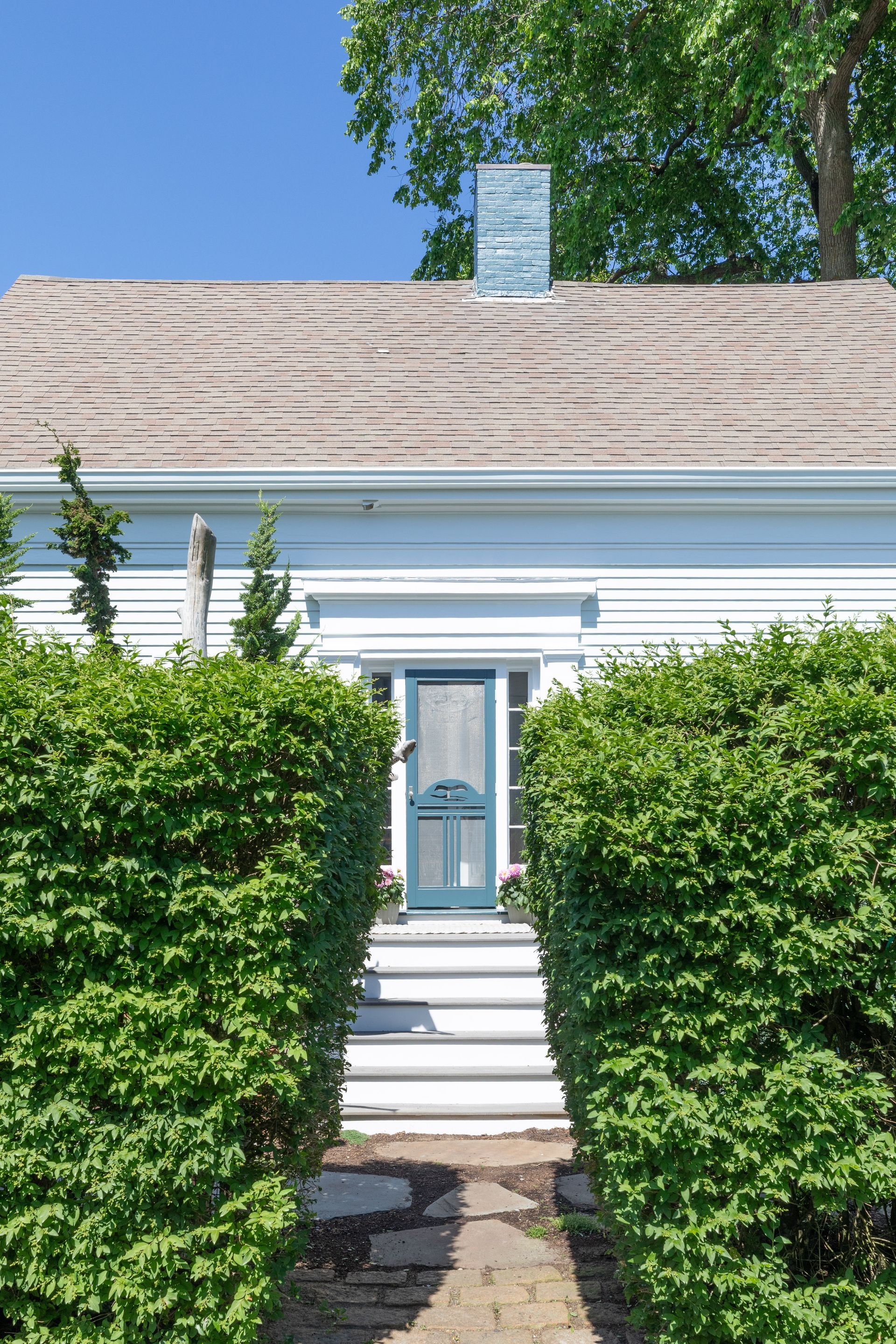 White house front with hedges, a narrow walkway, and a blue door under a shingled roof.