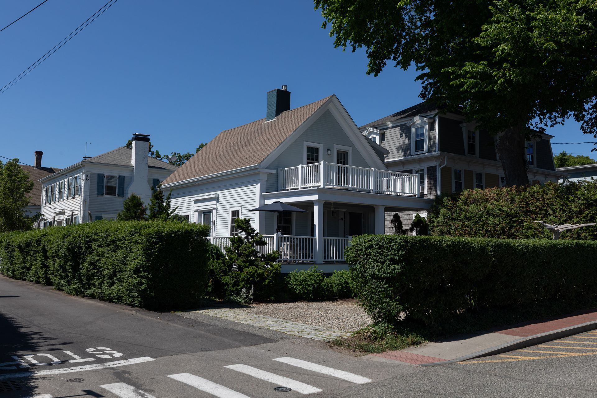 White house with balconies beside a hedge-lined street under a clear blue sky