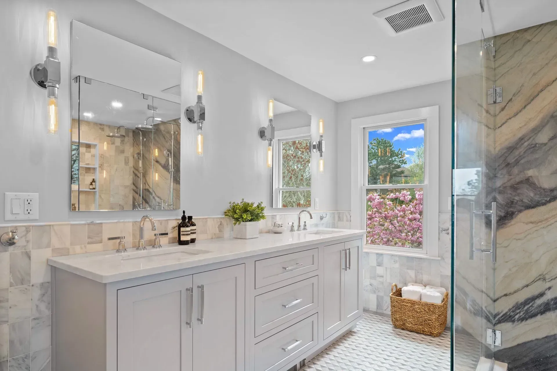 Bright modern bathroom with double vanity, large mirror, glass shower, and a window with a basket below.