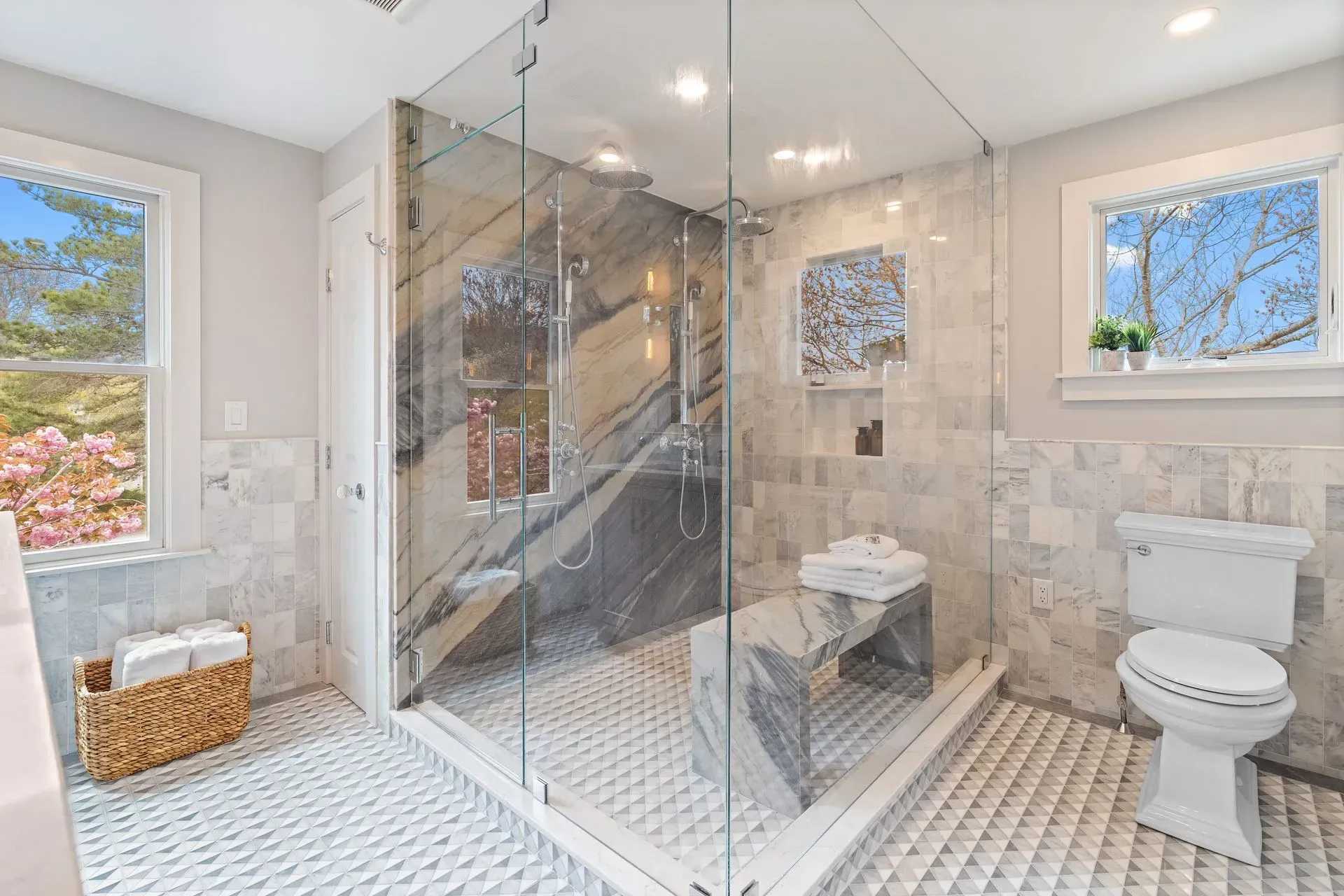 Bright tiled bathroom with glass shower, toilet, woven basket, and windows letting in natural light