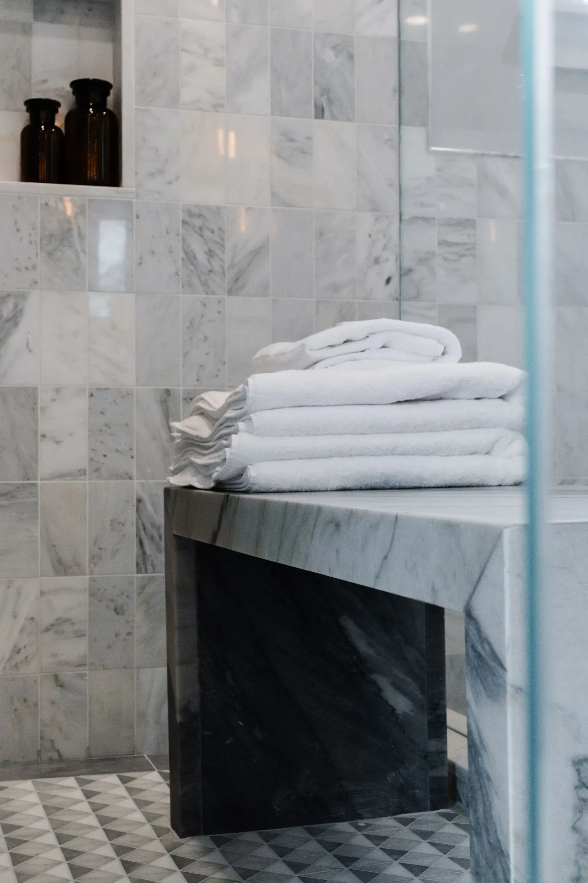 Bathroom with gray marble tiles, folded white towels on a dark bench, and a glass shower door