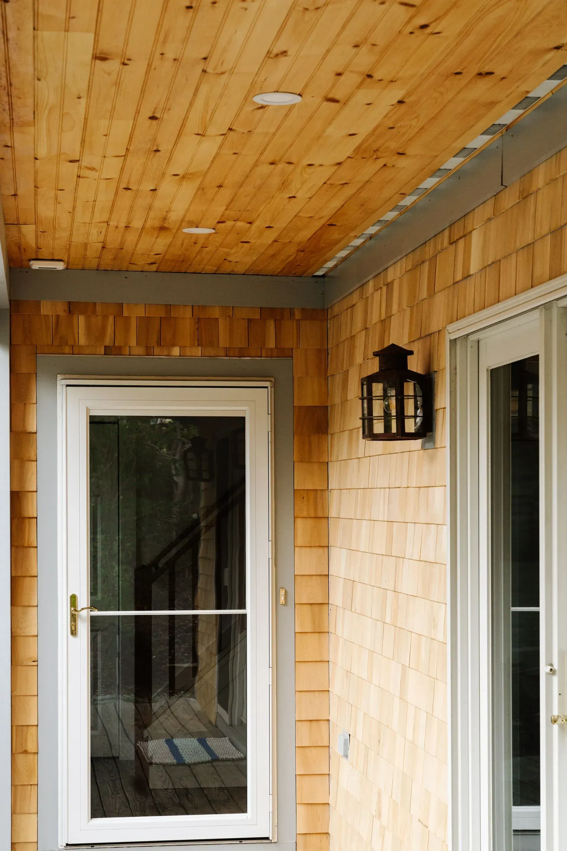 Front porch with wood-paneled ceiling, two white doors, and a black wall lantern.