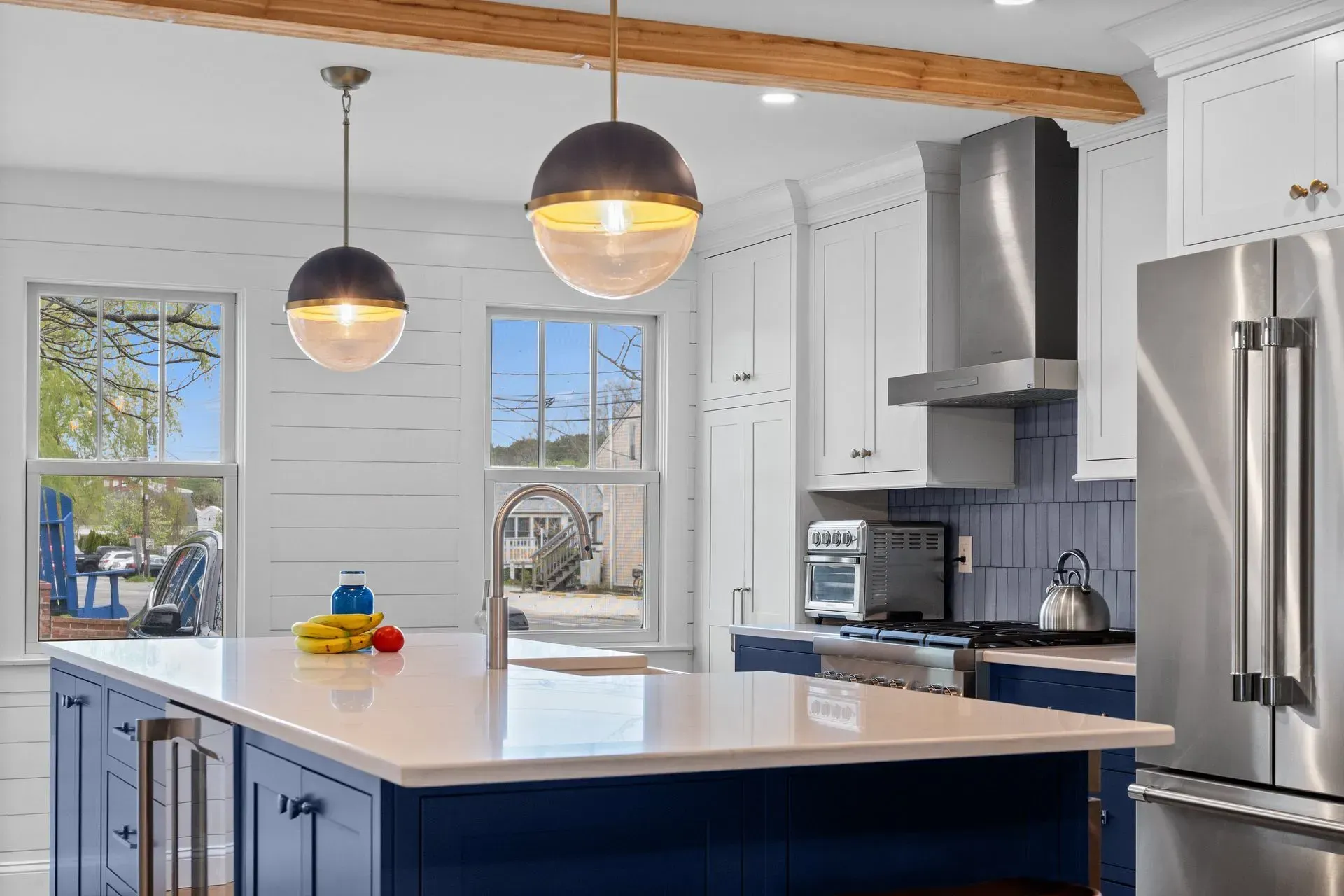 Bright modern kitchen with white island, blue cabinets, pendant lights, stainless appliances, and wood beam ceiling