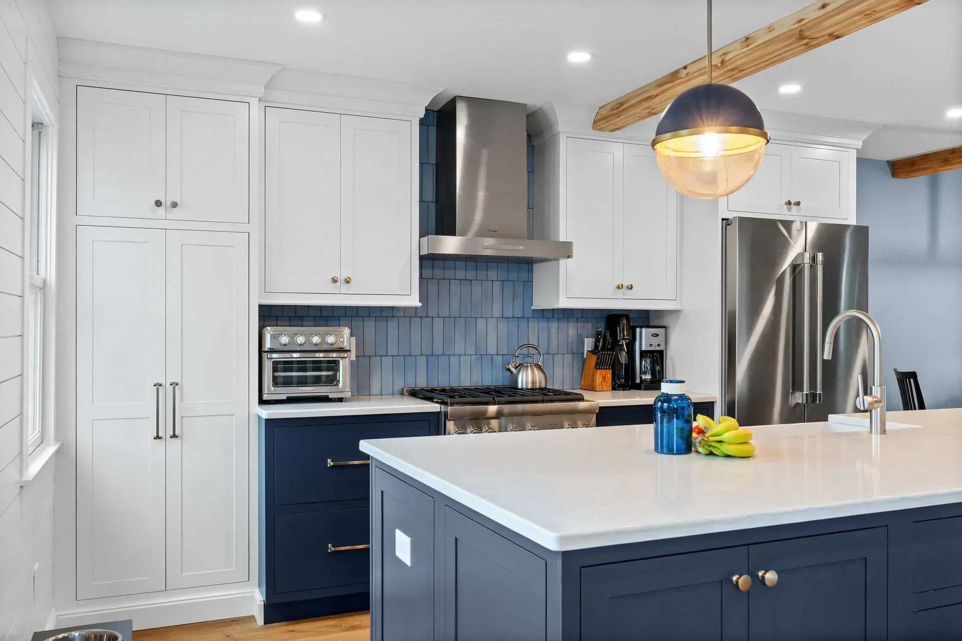 Modern white kitchen with navy island, stainless appliances, blue tile backsplash, and pendant light