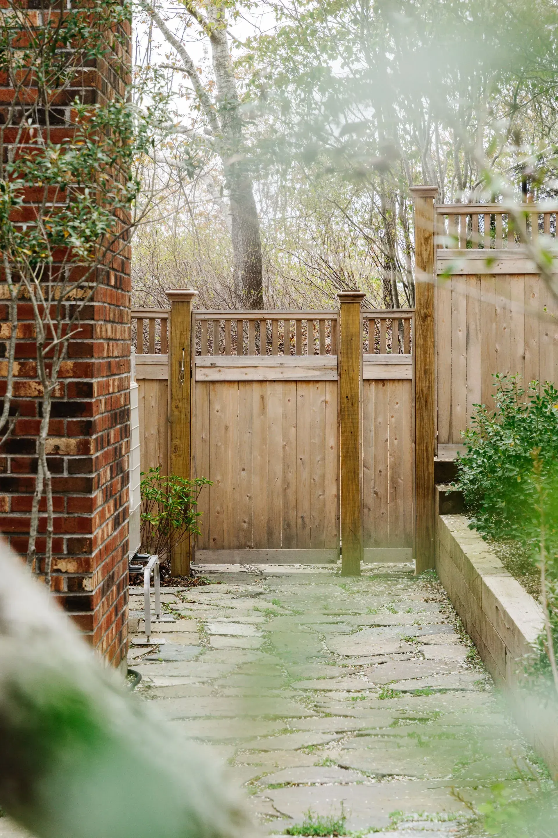 Narrow stone path leading to a wooden fence gate beside a brick wall and garden greenery.