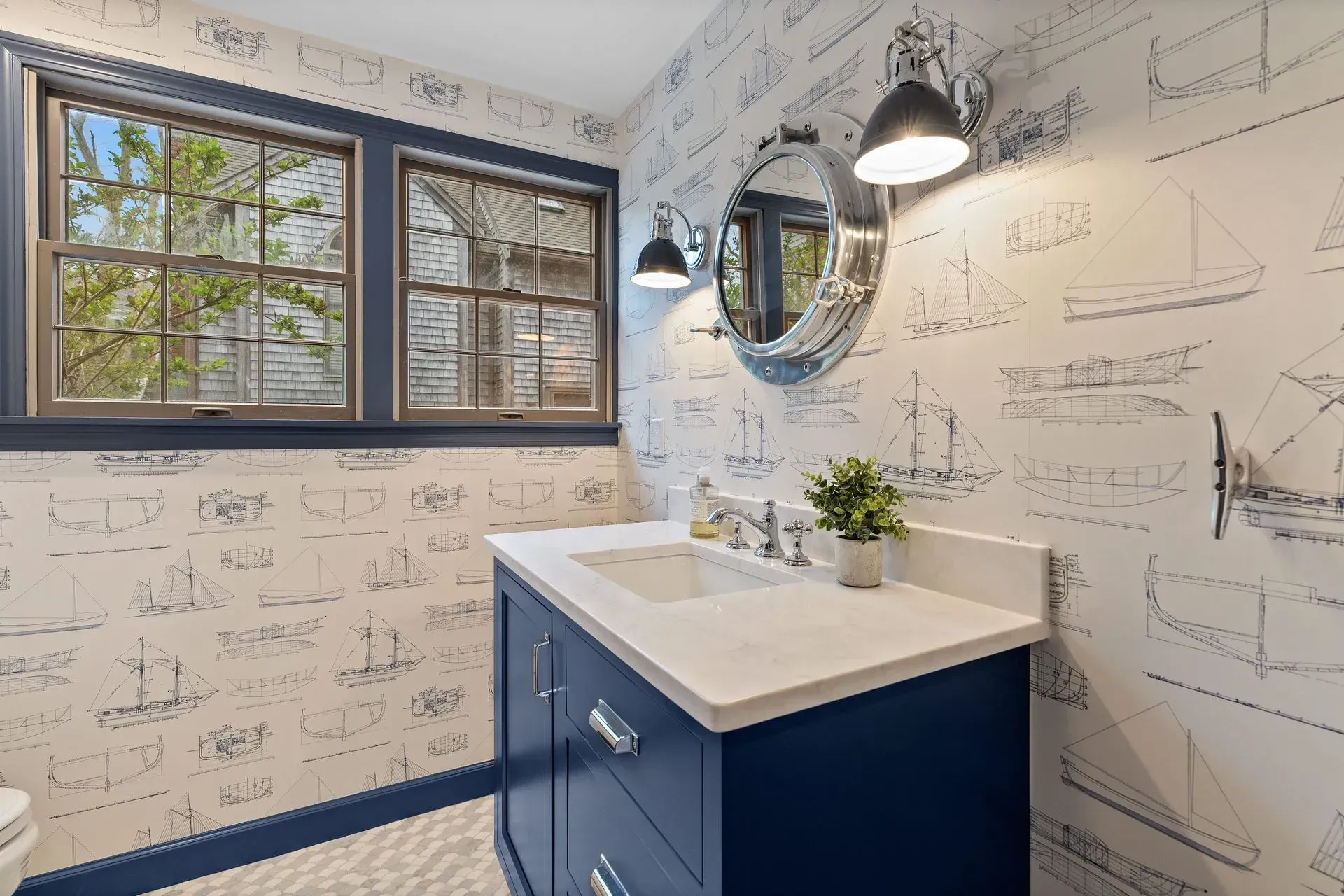 Blue vanity in a small bathroom with nautical wallpaper, window, and round mirror