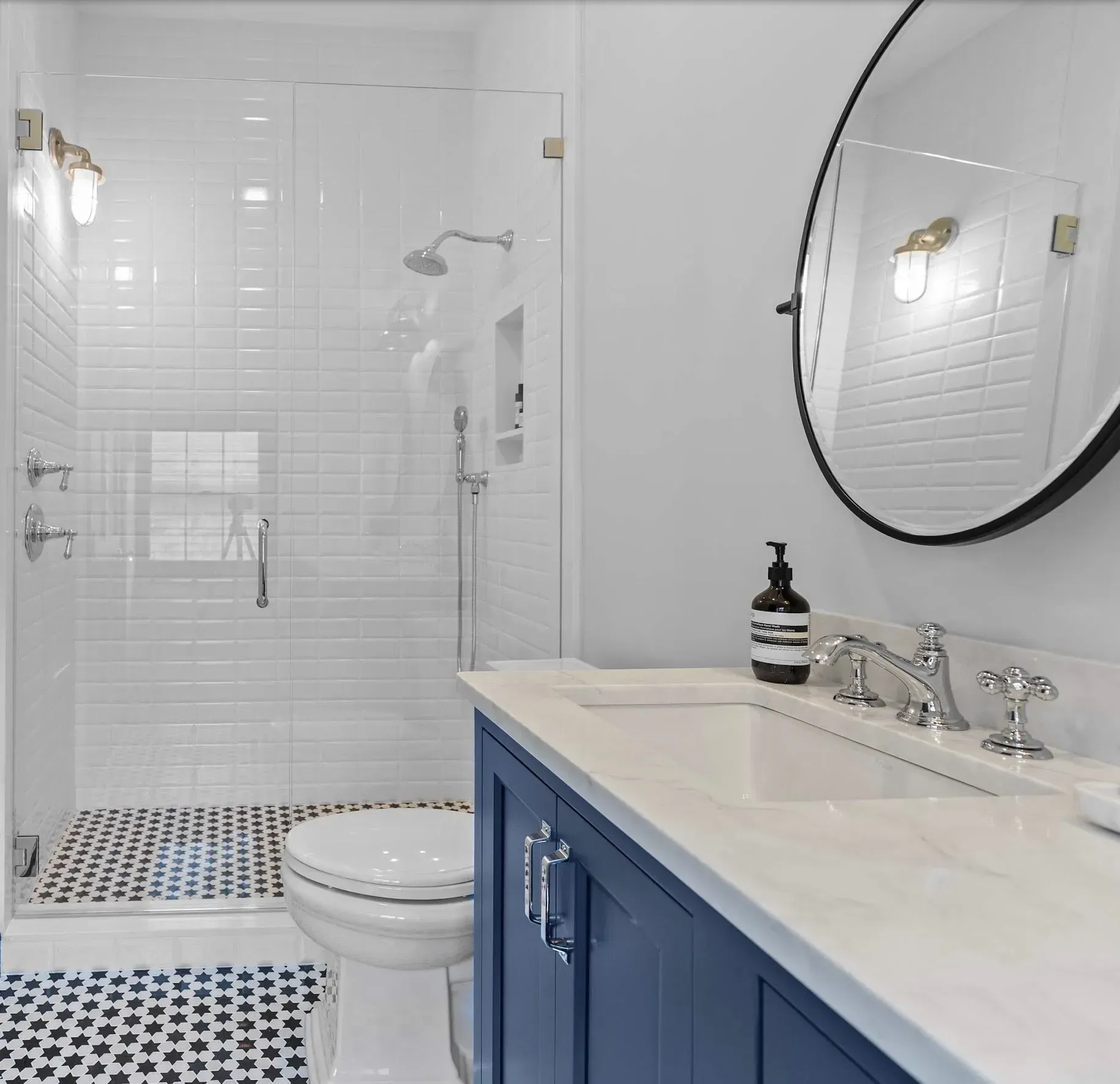 Bright white bathroom with blue vanity, round mirror, glass shower, and black-and-white tile floor.