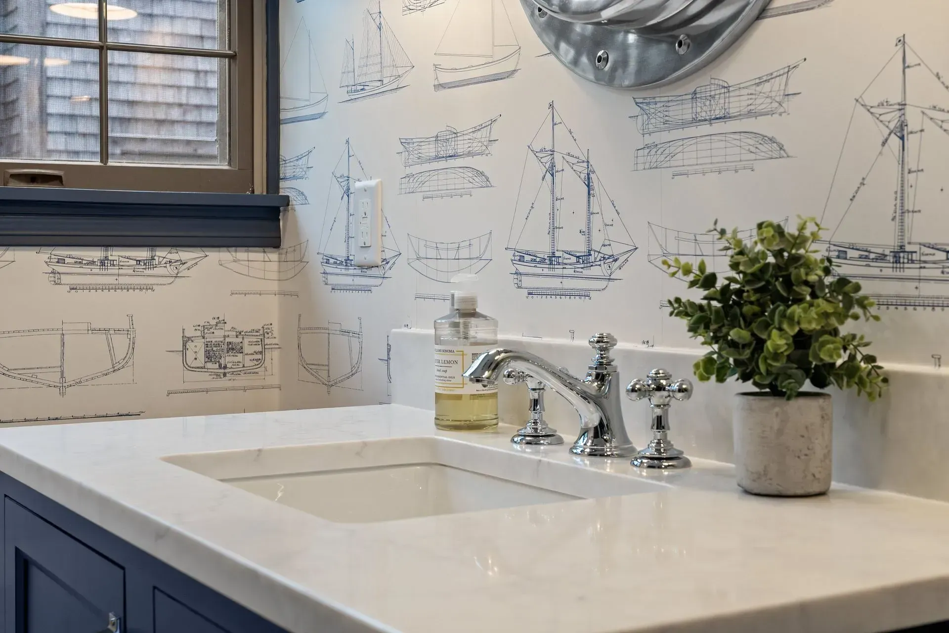 Modern bathroom sink with white countertop, chrome faucet, nautical wallpaper, and a potted plant.