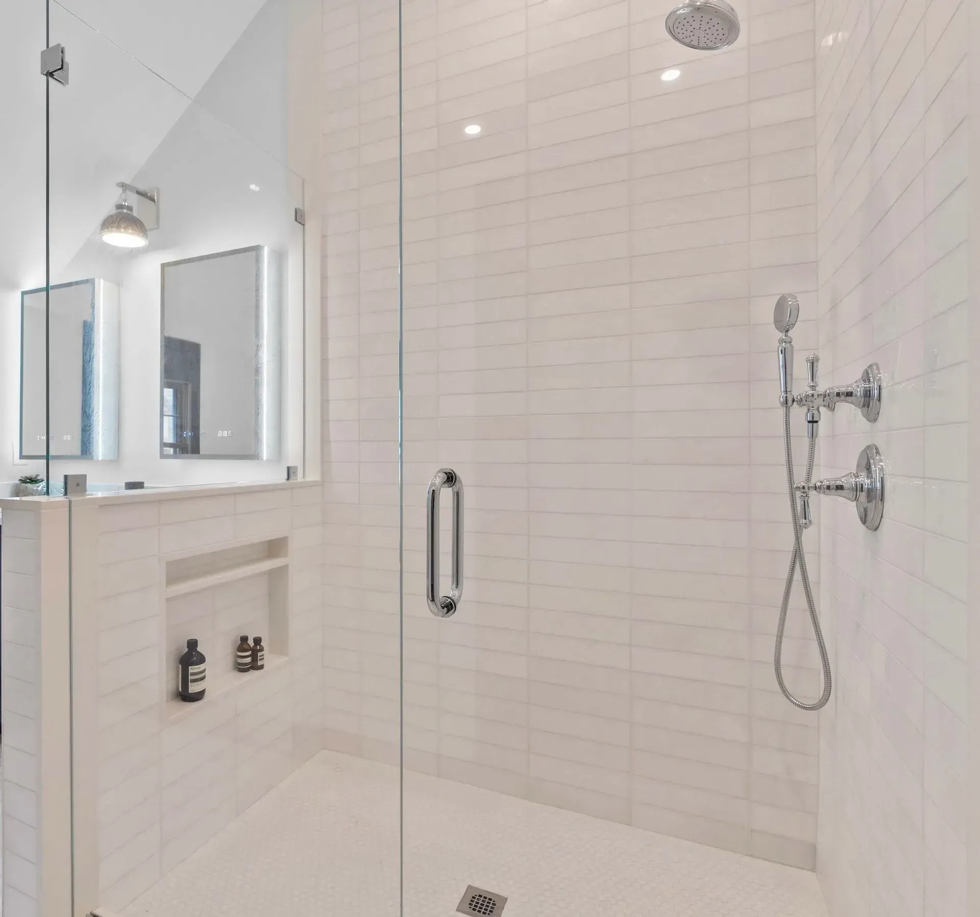 Modern white tiled shower with glass door, chrome fixtures, built-in niche, and vanity mirror visible outside.
