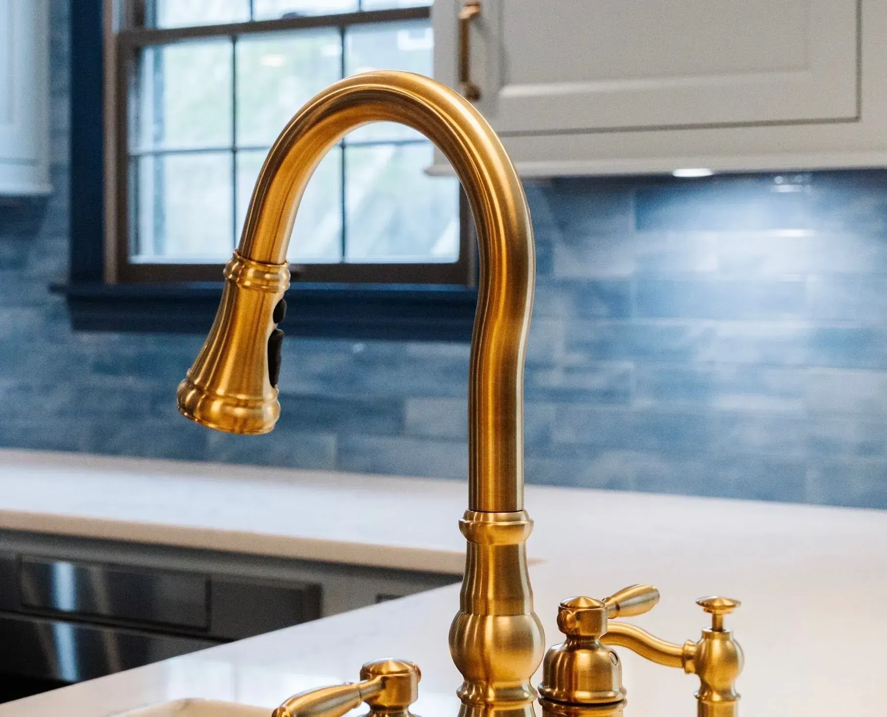 Golden kitchen faucet over a sink with a blue tile backsplash and white cabinets in the background