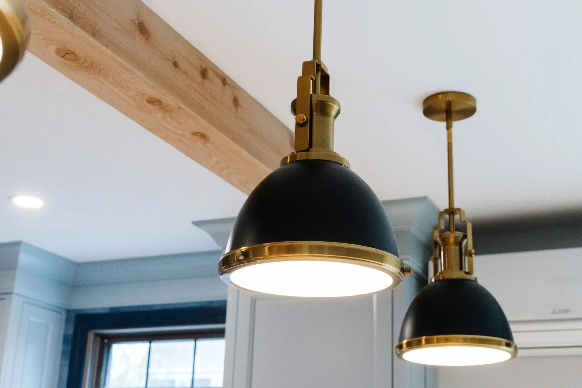 Two black-and-brass pendant lights hanging from a wooden beam in a bright kitchen