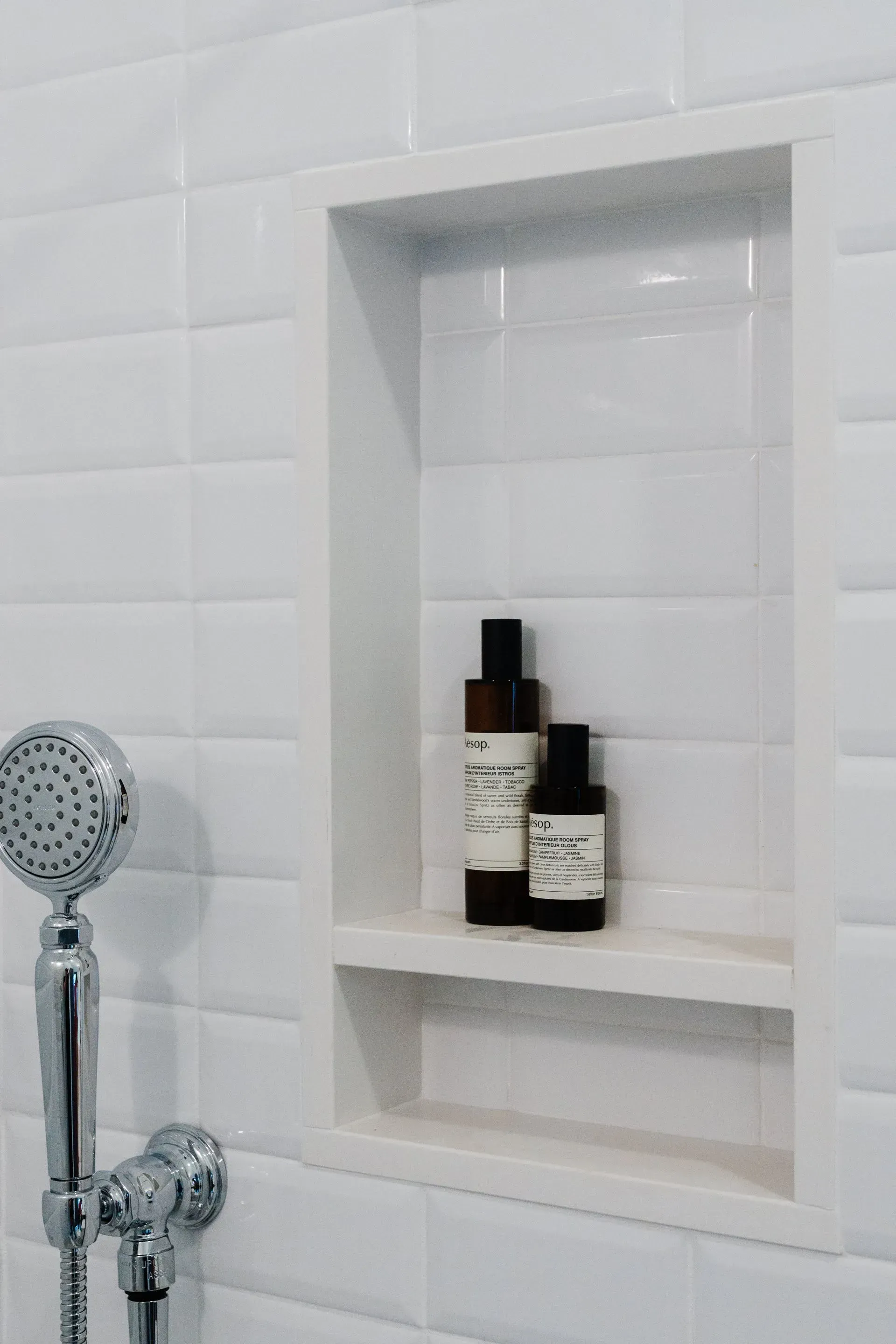 Shower niche with white tile, built-in shelf, and three toiletry bottles beside a handheld showerhead