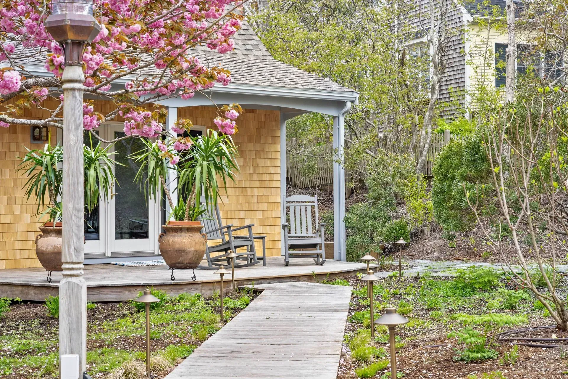 Garden path leading to a small porch with rocking chairs, potted plants, and pink blossoms