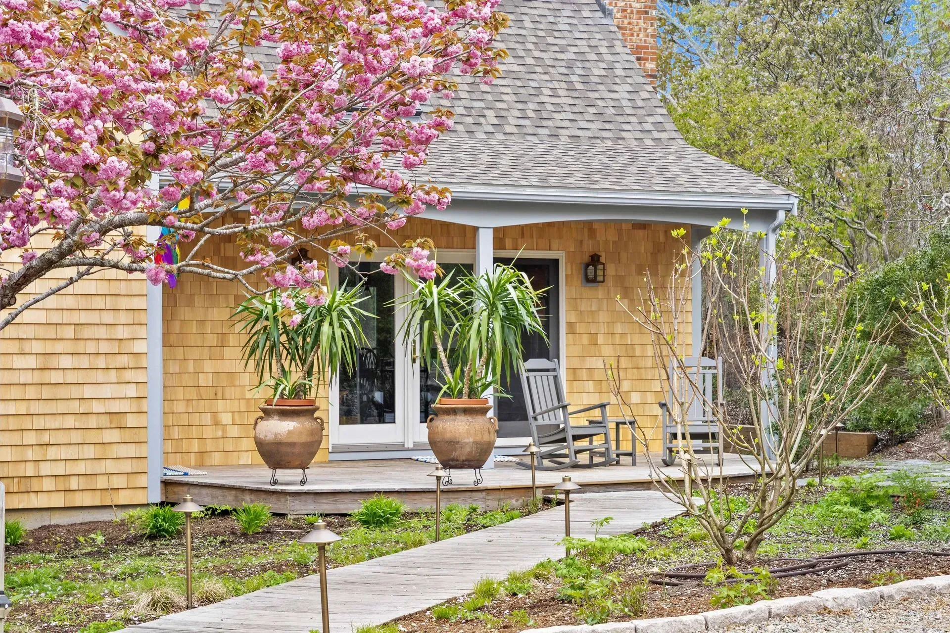 Cozy stone cottage porch with rocking chairs, potted plants, and a blooming pink tree in front